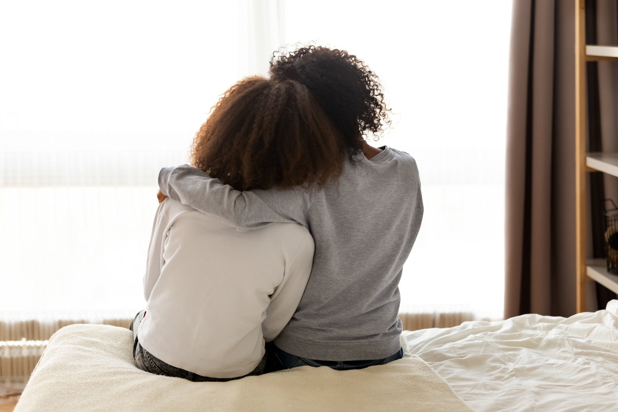 Zwei Frauen von hinten, die auf einem Bett sitzen und sich in den Arm nehmen