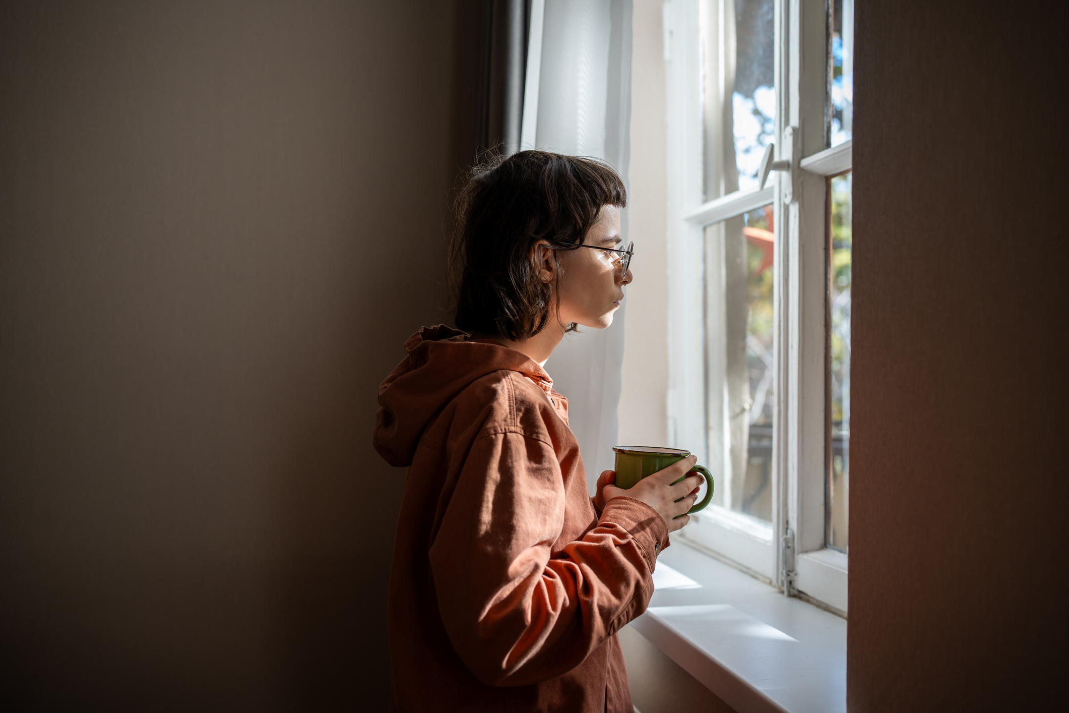Ein junger Mensch mit Brille und einer Tasse in der Hand steht vor einem Fenster und schaut nachdenklich heraus.