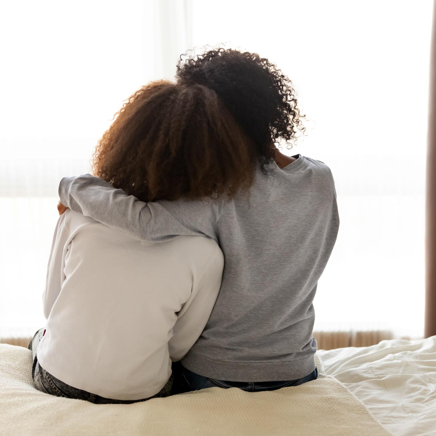 Zwei Frauen von hinten, die auf einem Bett sitzen und sich in den Arm nehmen
