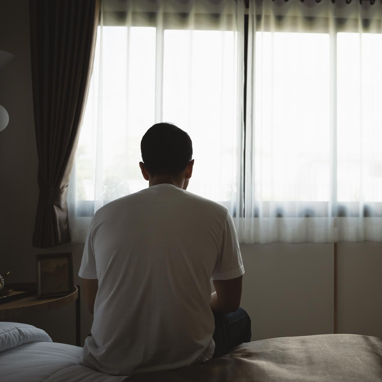Ein Mann sitzt mit dem Rücken zur Kamera auf einem Bett vor einem Fenster in einem dunklen Raum.