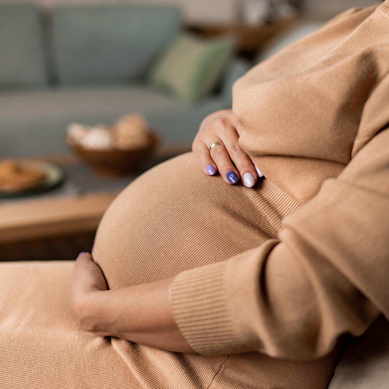 Eine Frau in einem beigen Kleid sitzt auf einem Stuhl und hält ihren Babybauch mit beiden Händen.