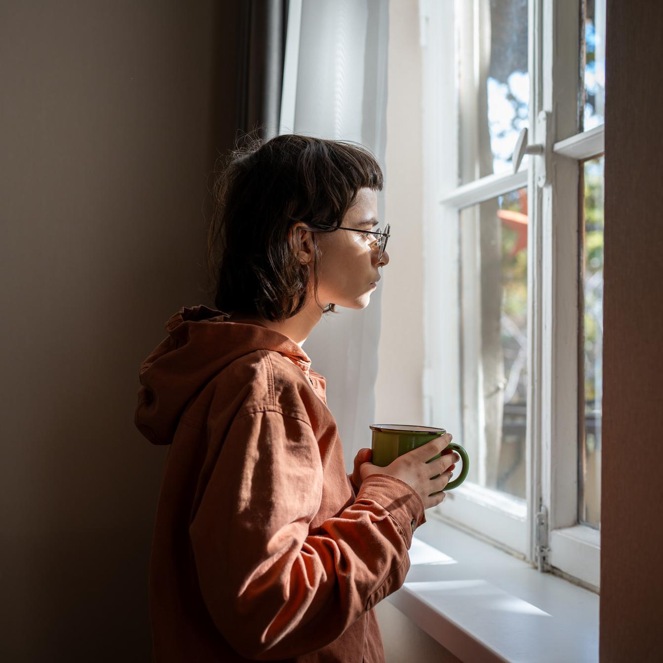 Ein junger Mensch mit Brille und einer Tasse in der Hand steht vor einem Fenster und schaut nachdenklich heraus.