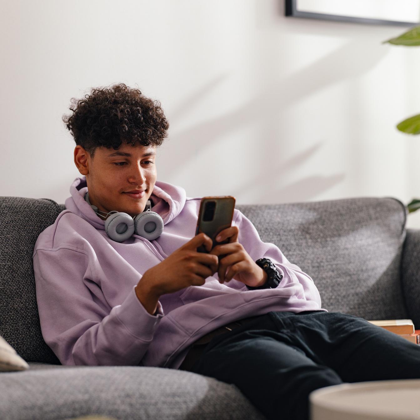 Ein Junge sitzt entspannt auf einem Sofa und schaut lächelnd in sein Smartphone